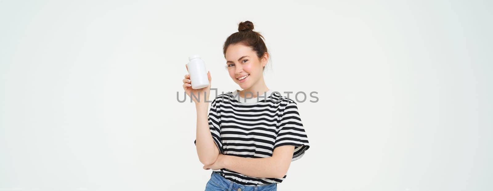 Young smiling woman recommends vitamins, shows bottle with dietary supplements, looks healthy and happy, isolated over white background.