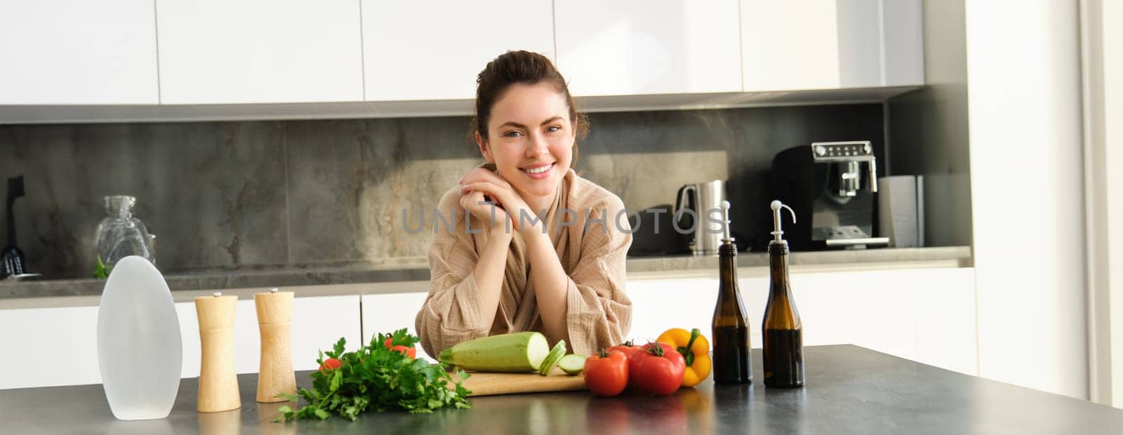 Portrait of smiling young woman cooking in kitchen in cosy clothes, standing near counter with chopping board, vegetables, zucchini, preparing dinner for family, making salad.