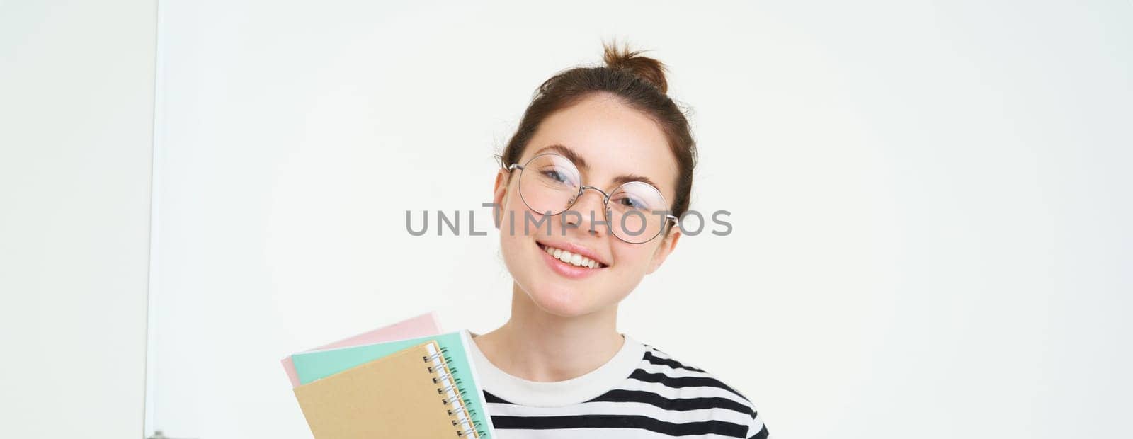 Portrait of young woman, tutor in glasses, holding her notebooks and documents, smiling at camera, standing over white background.