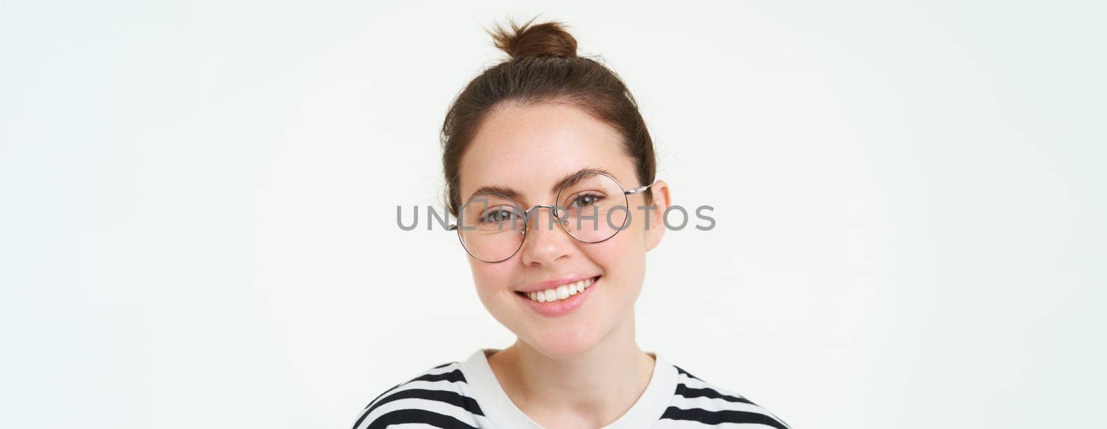 Close up portrait of cute young woman in glasses, looking at camera and smiling, trying on new spectacles, standing over white background.