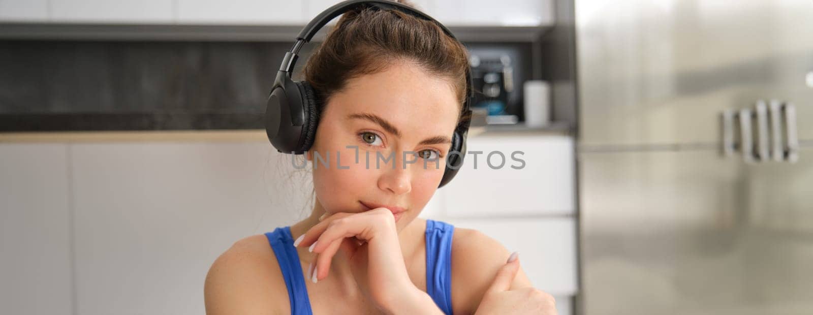 Close up portrait of brunette fitness girl, listens music in wireless headphones, smiles at camera, does workout alone at home, sits on floor and exercises by Benzoix