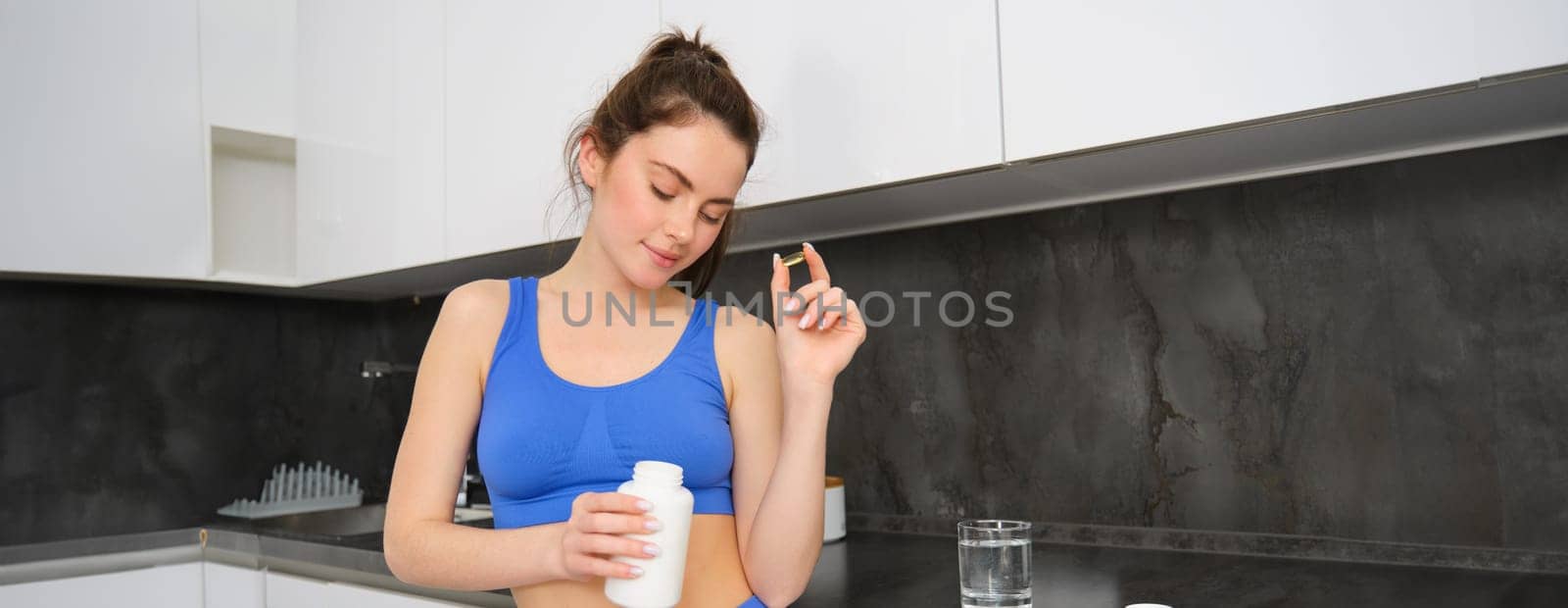 Image of young brunette woman with slim, fit body, holding dietary supplements, vitamin B, fish oil, standing in kitchen.