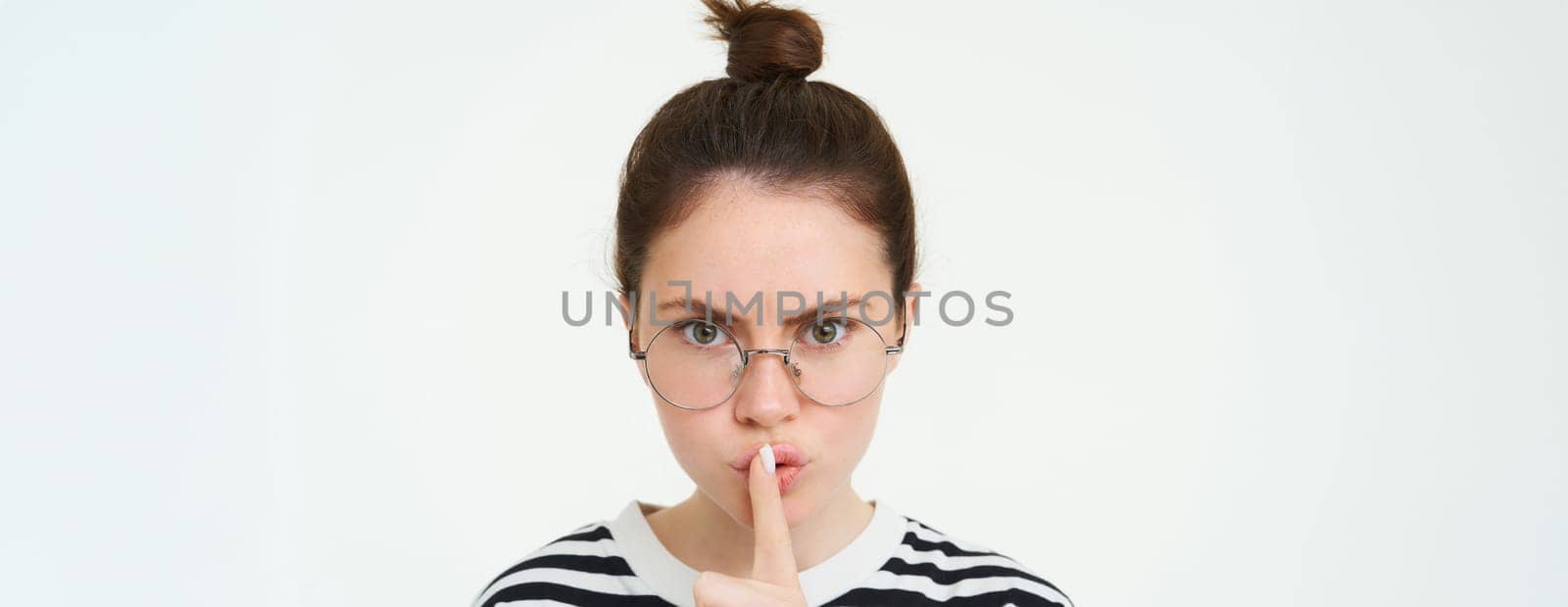 Image of serious woman in glasses, teacher shushing, makes shush, hush gesture, press finger to lips and frowning from disapproval, standing over white background by Benzoix