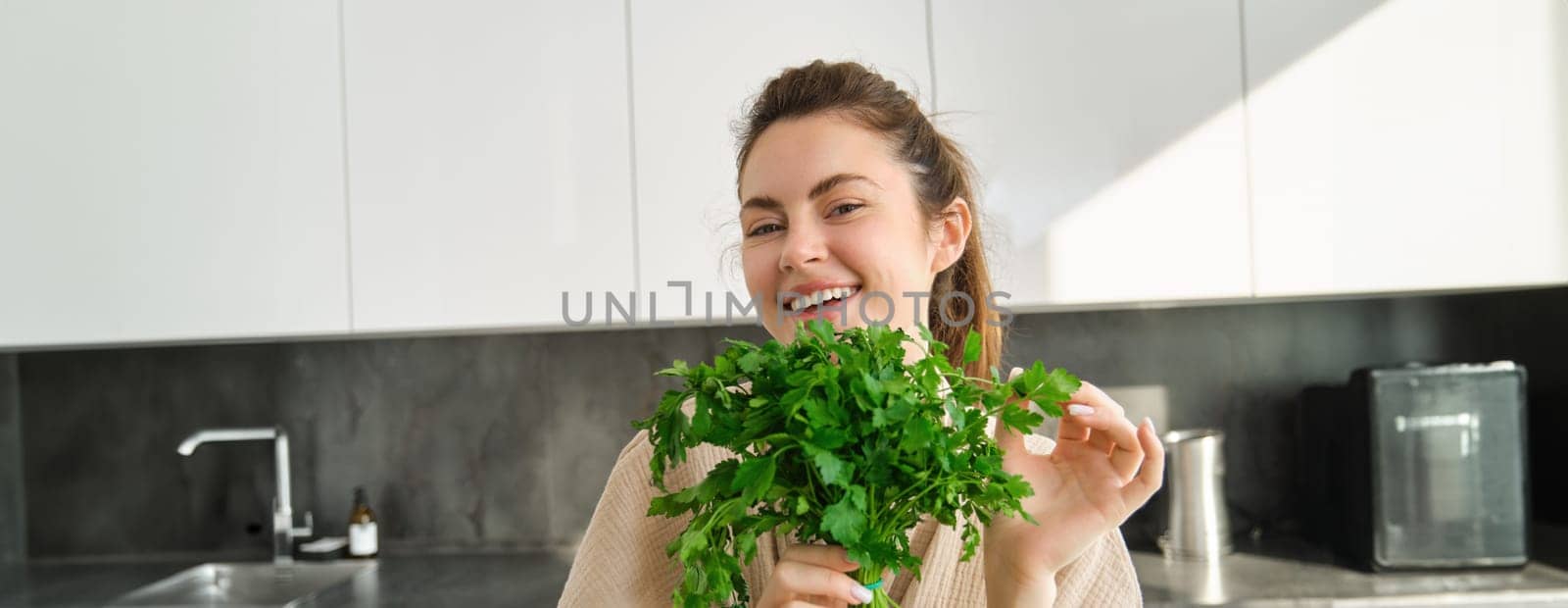 Portrait of beautiful smiling girl with bouquet of parsley, standing in the kitchen and cooking, adding herbs to healthy fresh salad or meal, preparing food.