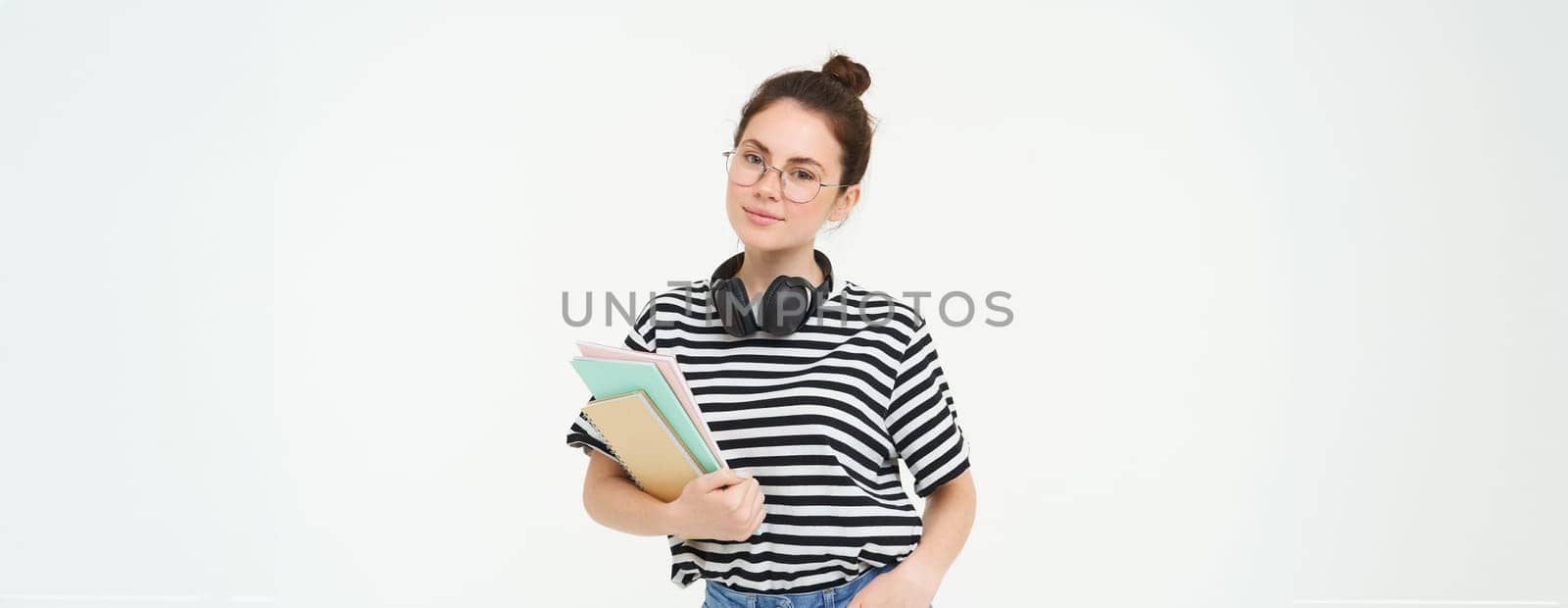 Education concept. Smiling brunette girl, student in casual clothes, holds her books, study material, wears headphones over neck, looks confident and happy, isolated over white background.