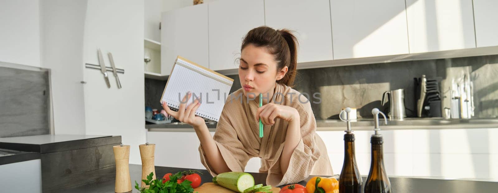 Portrait of woman checking grocery list, looking at vegetables, holding notebook, reading recipe while cooking meal in the kitchen, chopping tomatoes and zucchini.