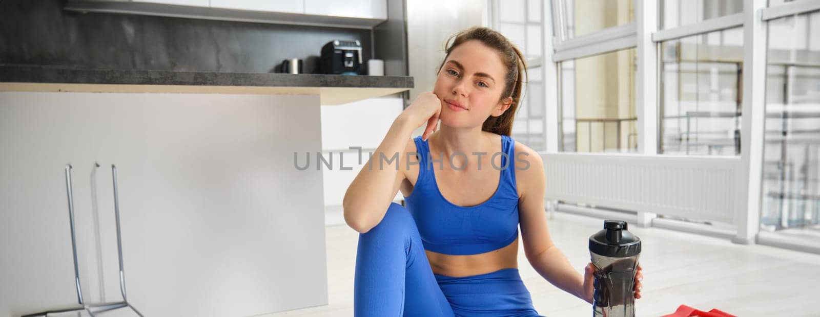 Smiling young woman doing sports in her house, looking at camera and smiling, drinking water after home fitness session.