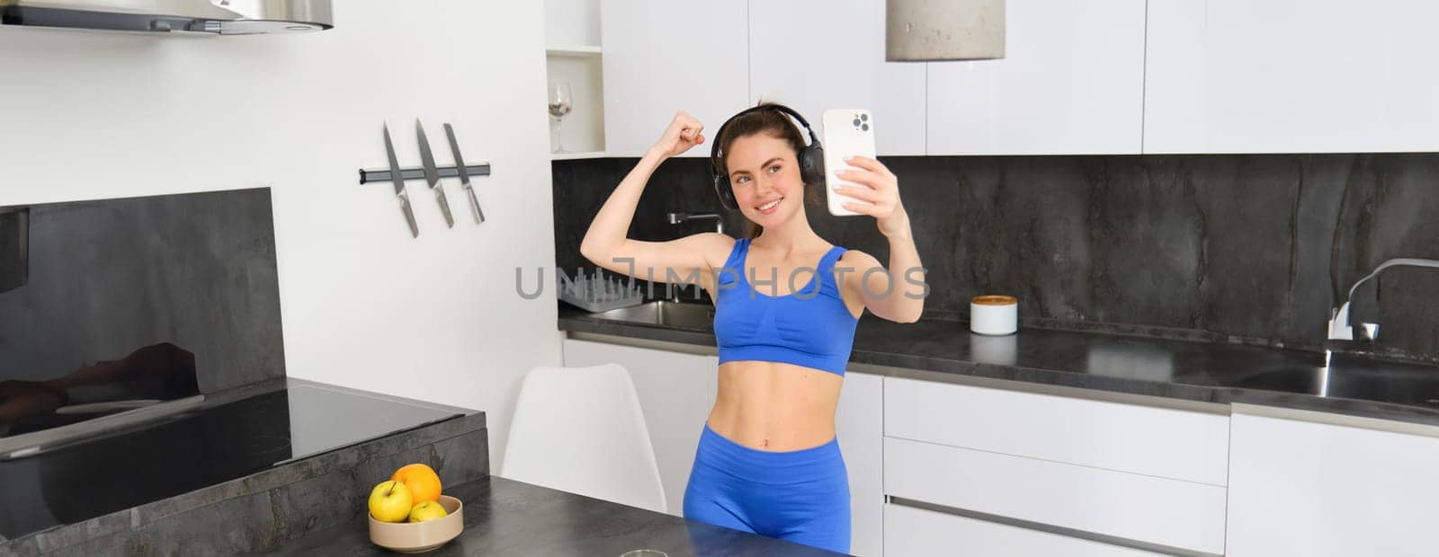 Portrait of beautiful young woman blogger, taking selfie in her workout clothes, shows her muscles, flexing biceps on smartphone camera, records video for social media by Benzoix
