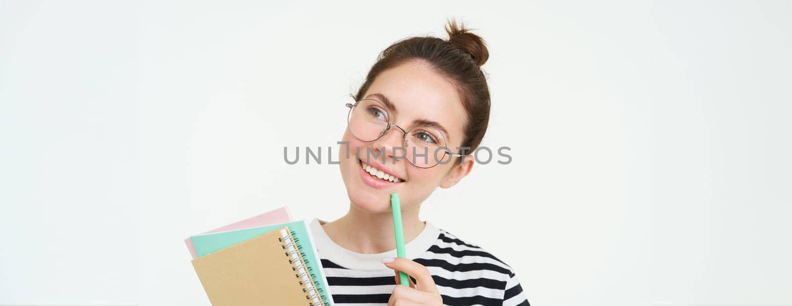 Portrait of smart girl in glasses, tutor holding pen and notebooks, student carry her homework notes, standing over white background.