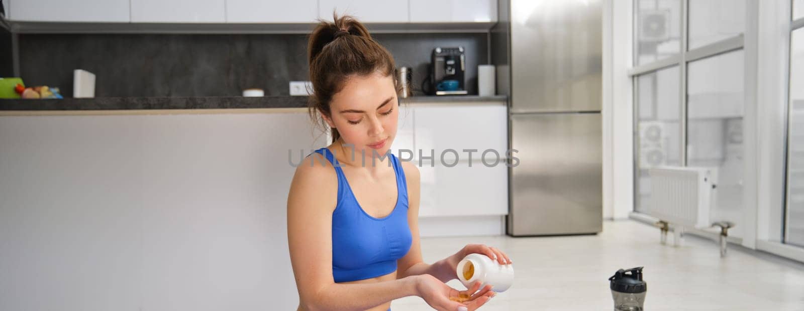 Portrait of beautiful and healthy fitness woman, taking vitamins, dietary supplements for muscles and bones, doing workout training at home by Benzoix