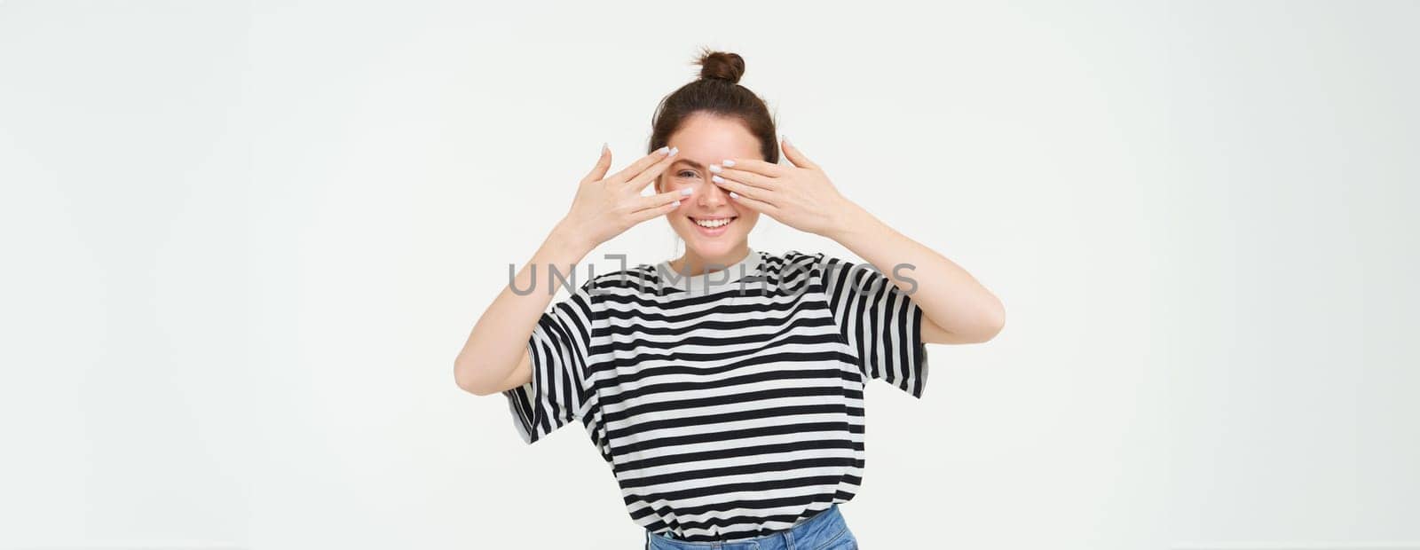 Portrait of girl waiting for surprise, covers eyes with hands, peeking through fingers, stands over white background.