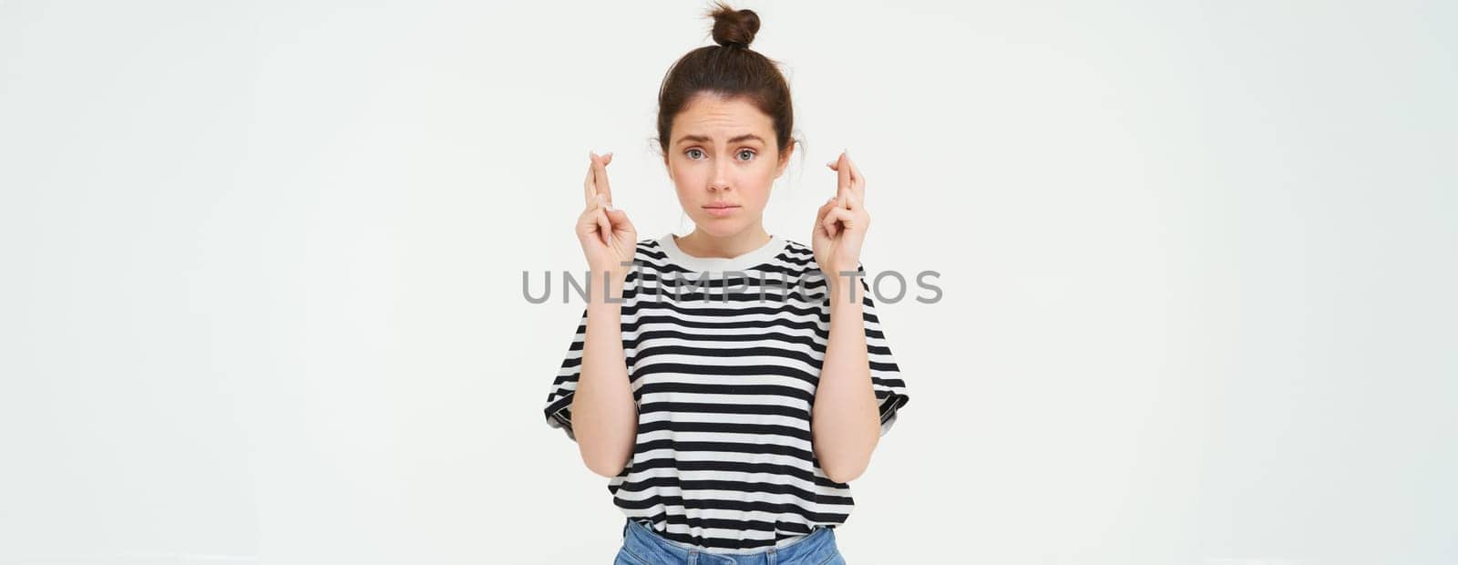 High hopes. Young woman cross fingers, makes wish, anticipates something, stands over white background.