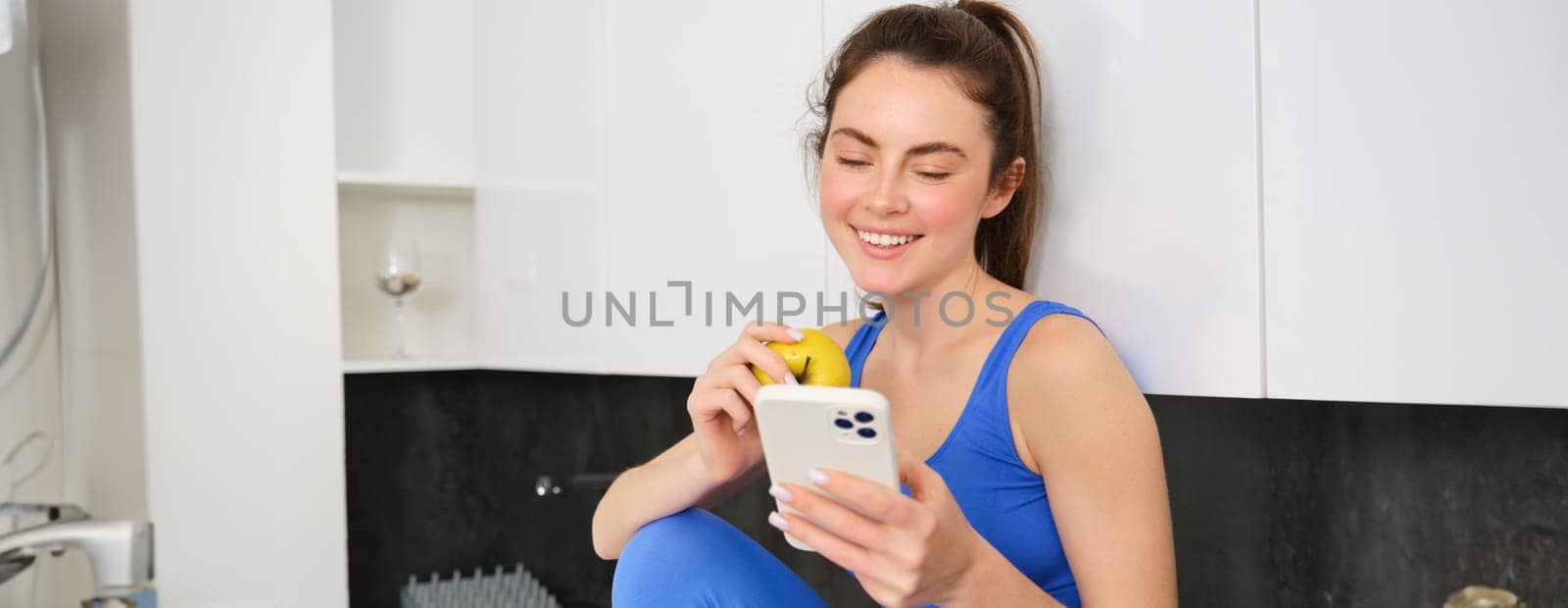 Healthy lifestyle and technology. Young beautiful girl, fitness instructor, looking at smartphone, reading on mobile phone and eating an apple, smiling happily.