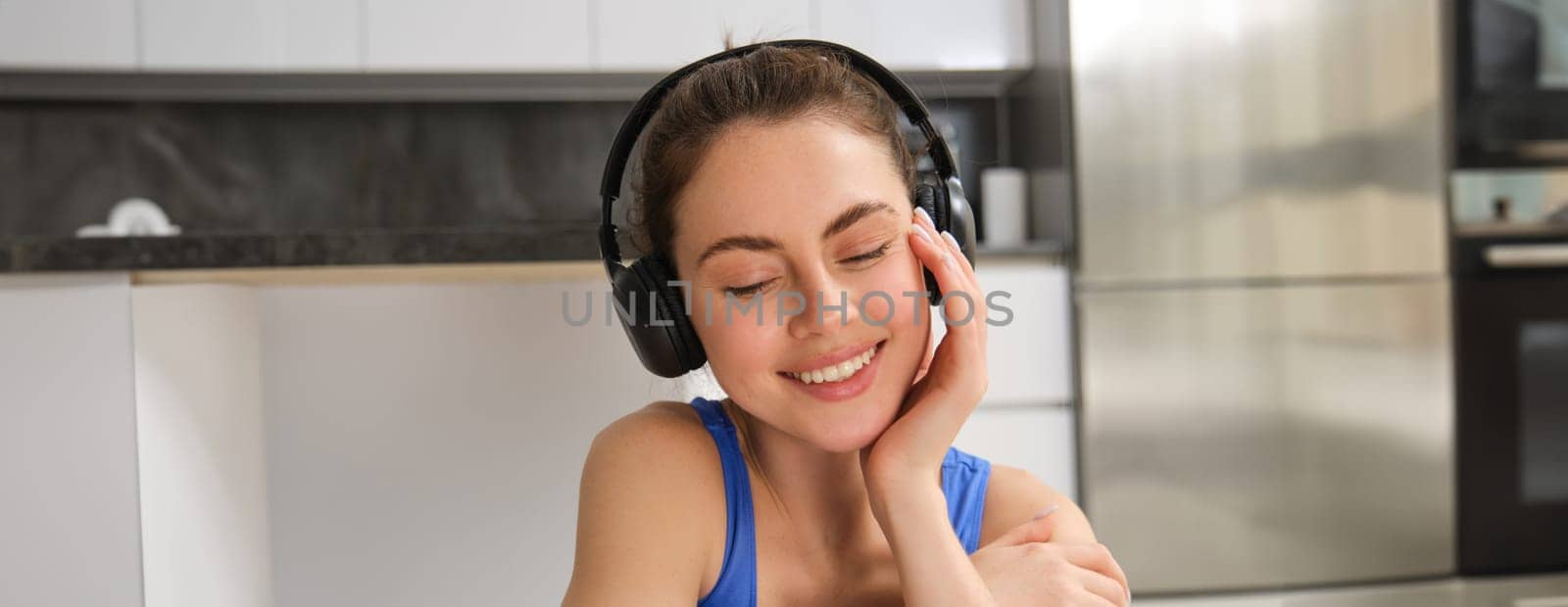 Close up portrait of beautiful brunette girl in sportswear, sits in wireless headphones and listens music, while doing workout training at home by Benzoix