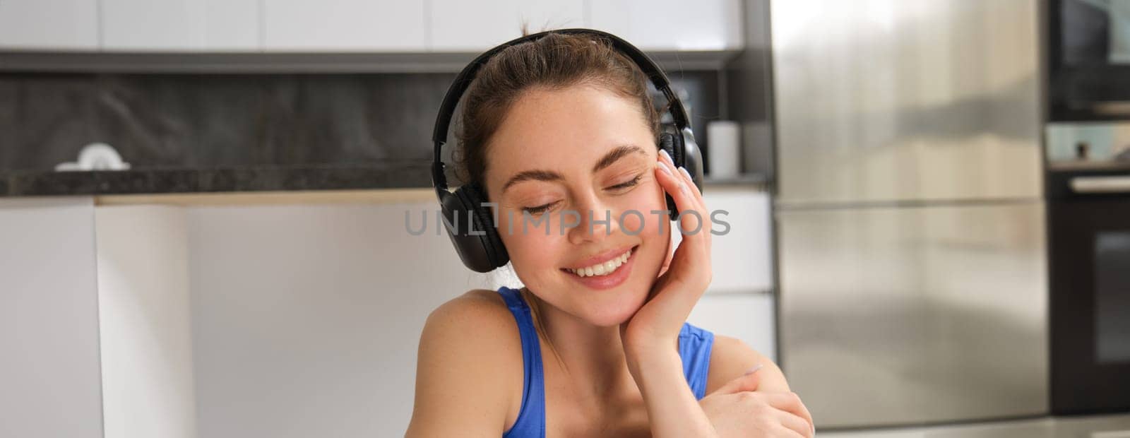 Close up portrait of beautiful brunette girl in sportswear, sits in wireless headphones and listens music, while doing workout training at home.