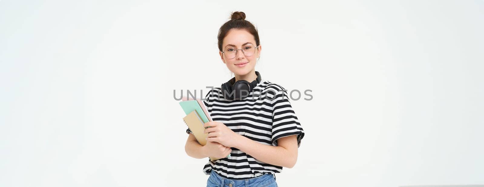 Young woman with notebooks, books and study material, posing over white background, wears headphones over neck by Benzoix