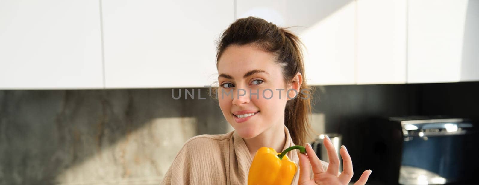 Close up portrait of beautiful woman smiling, showing yellow pepper, vegetarian girl making dinner, preparing meal with vegetables, posing in kitchen.