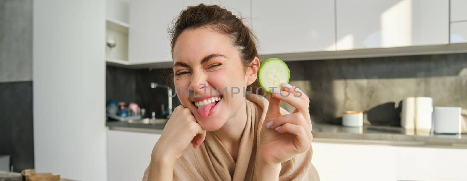 Portrait of beautiful brunette girl cooking in the kitchen, posing in bathrobe at home, holding zucchini, showing happy smile, making healthy food, vegetarian meal by Benzoix