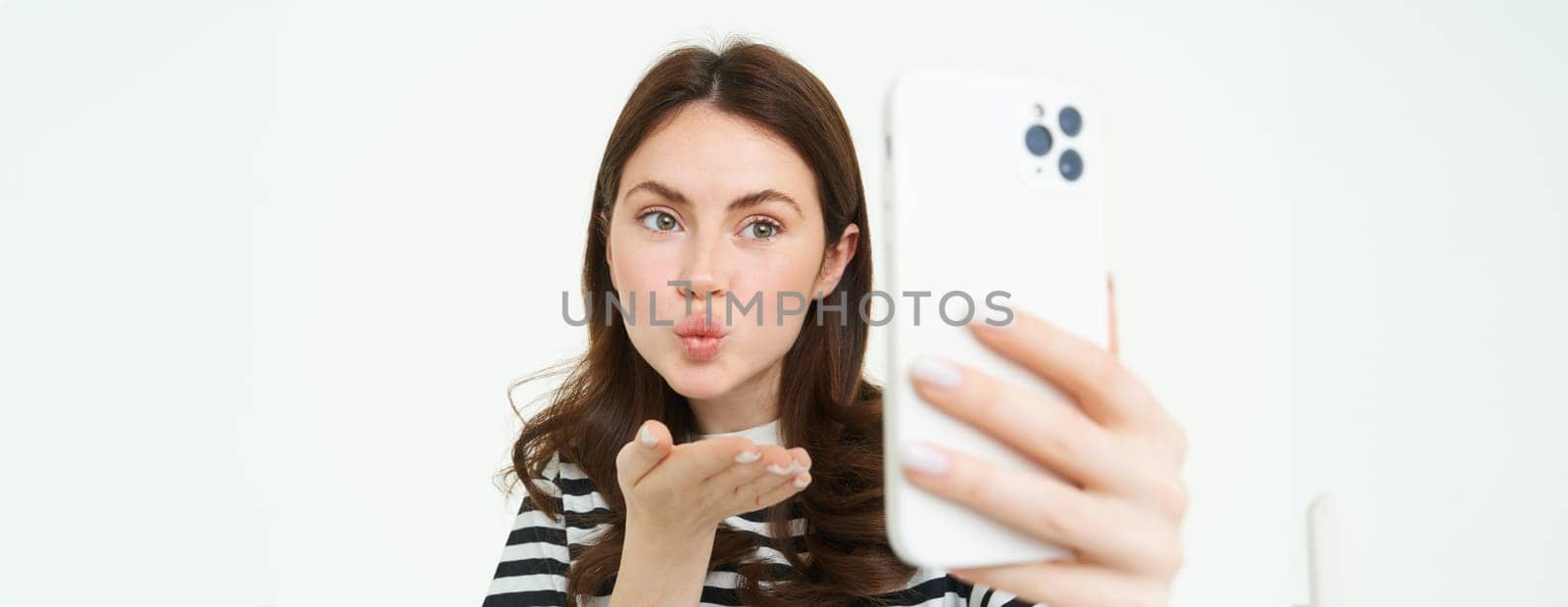 Portrait of young european woman taking selfie on smartphone, holding white mobile phone and posing for photos, isolated against white background by Benzoix
