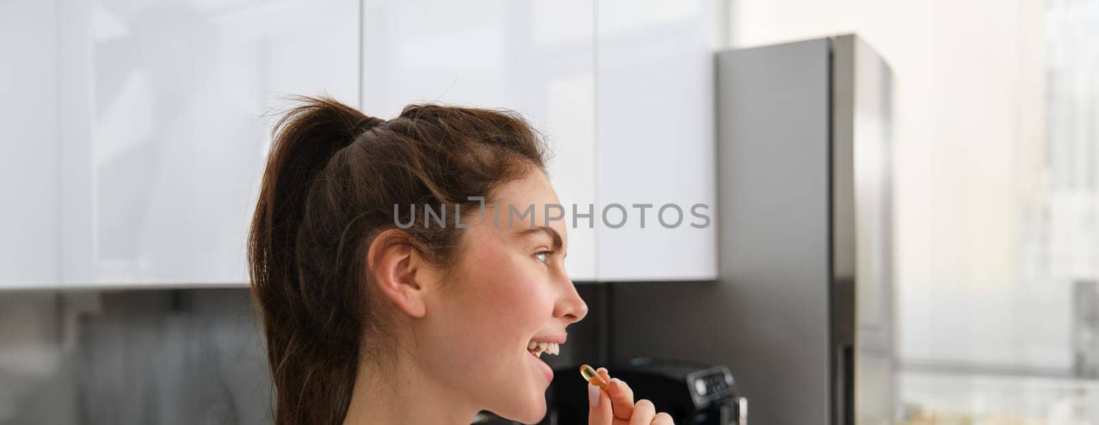 Sport and healthcare. Close up portrait of woman taking supplements, buds, holding vitamin tablet near mouth, using fish oil for healthy strong body, standing in kitchen.