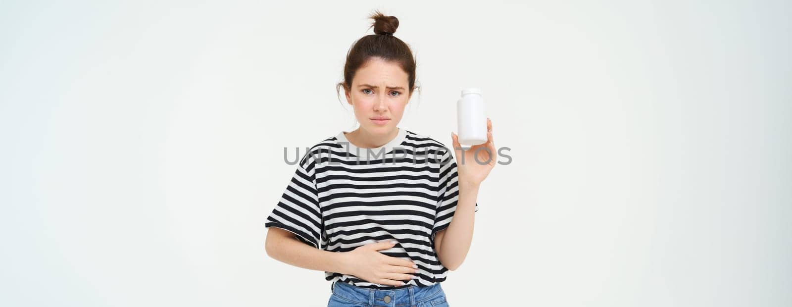 Woman takes painkillers, pills from stomach ache, menstrual pain, holds hand on belly, shows bottle with tablets, stands over white background by Benzoix