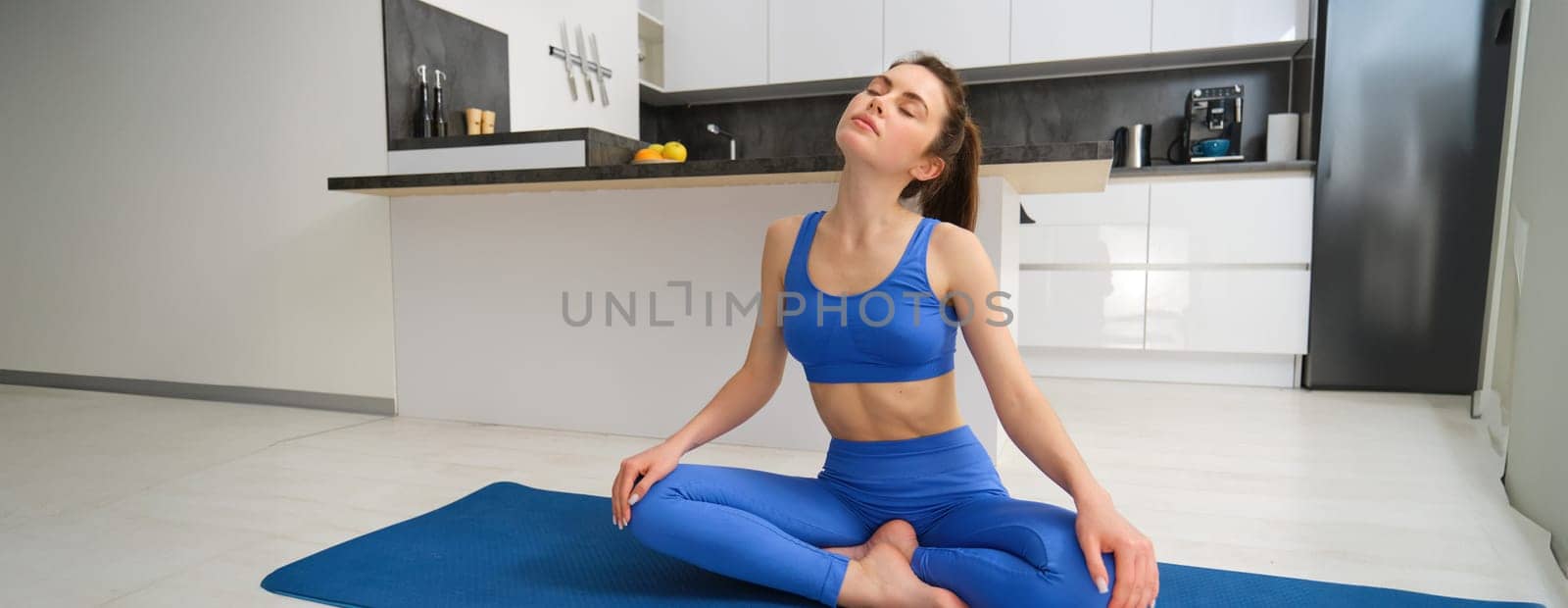 Portrait of young sportswoman, stretching her neck, warm-up before yoga exercises, doing fitness workout on rubber mat, wearing blue sportsbra and leggings by Benzoix