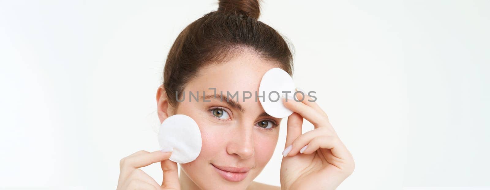 Portrait of young woman cleansing her face, takes off her makeup, washing her face, using skincare routine with cotton pads, isolated over white background by Benzoix