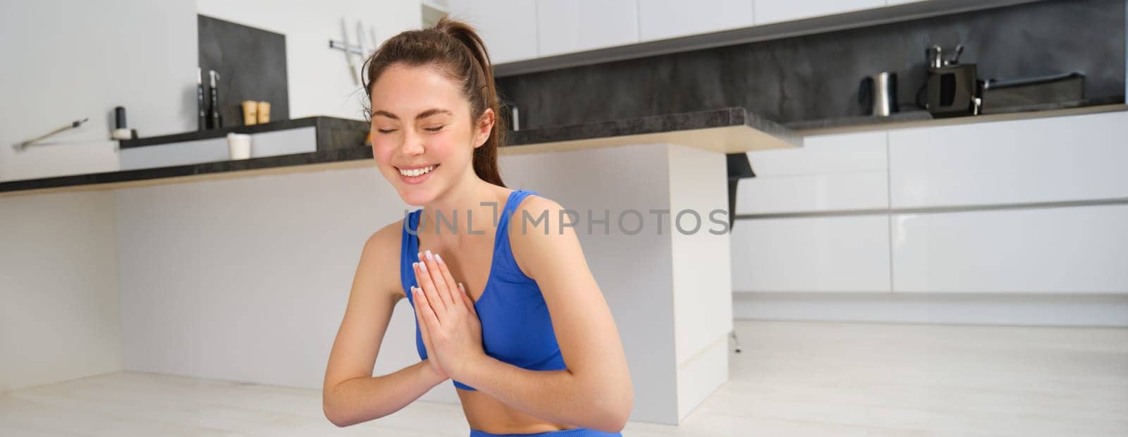 Young healthy beautiful woman in sports top and leggings practicing yoga at home, sitting in lotus position on yoga mat, meditating, smiling, relaxed, eyes closed, meditation concept by Benzoix