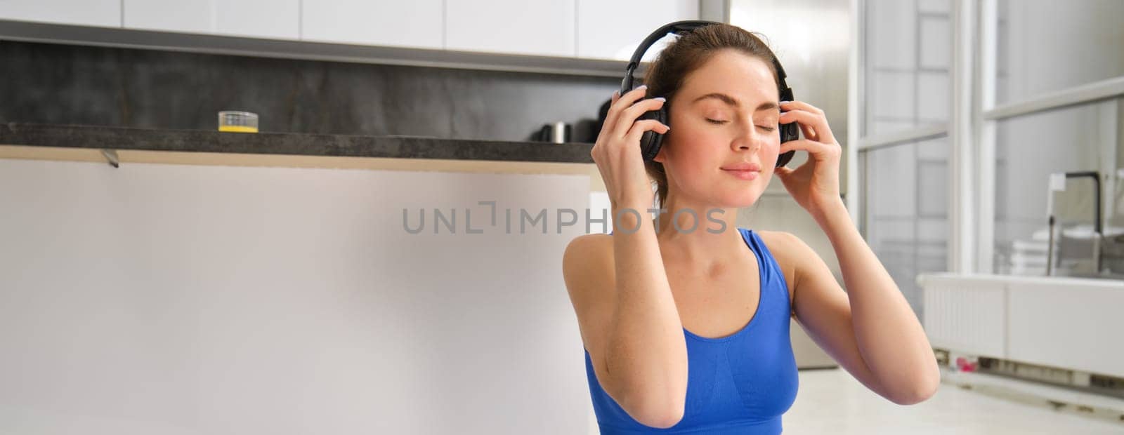 Smiling fitness woman puts on wireless headphones before her home workout, yoga training in blue tracksuit by Benzoix