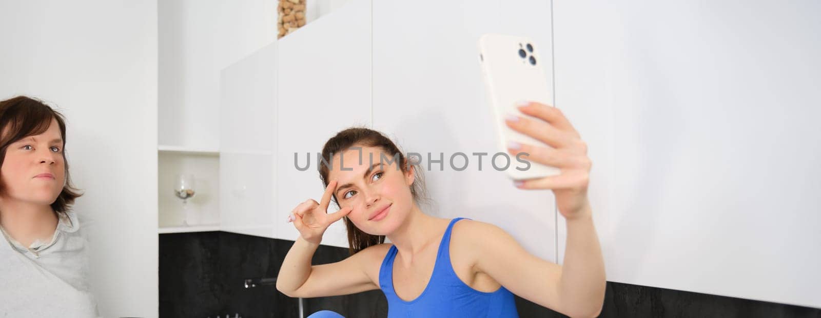 Portrait of smiling, beautiful fitness instructor, posing for fitness blog, selfie on smartphone, sitting in kitchen, looking happy.