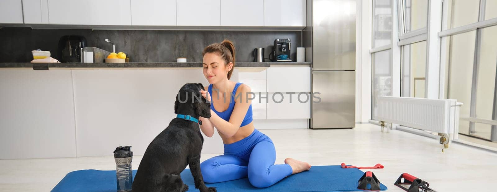 Image of young woman does workout from home, sits on yoga mat in living room and plays with her black dog, puppy distracts girl from doing exercises.