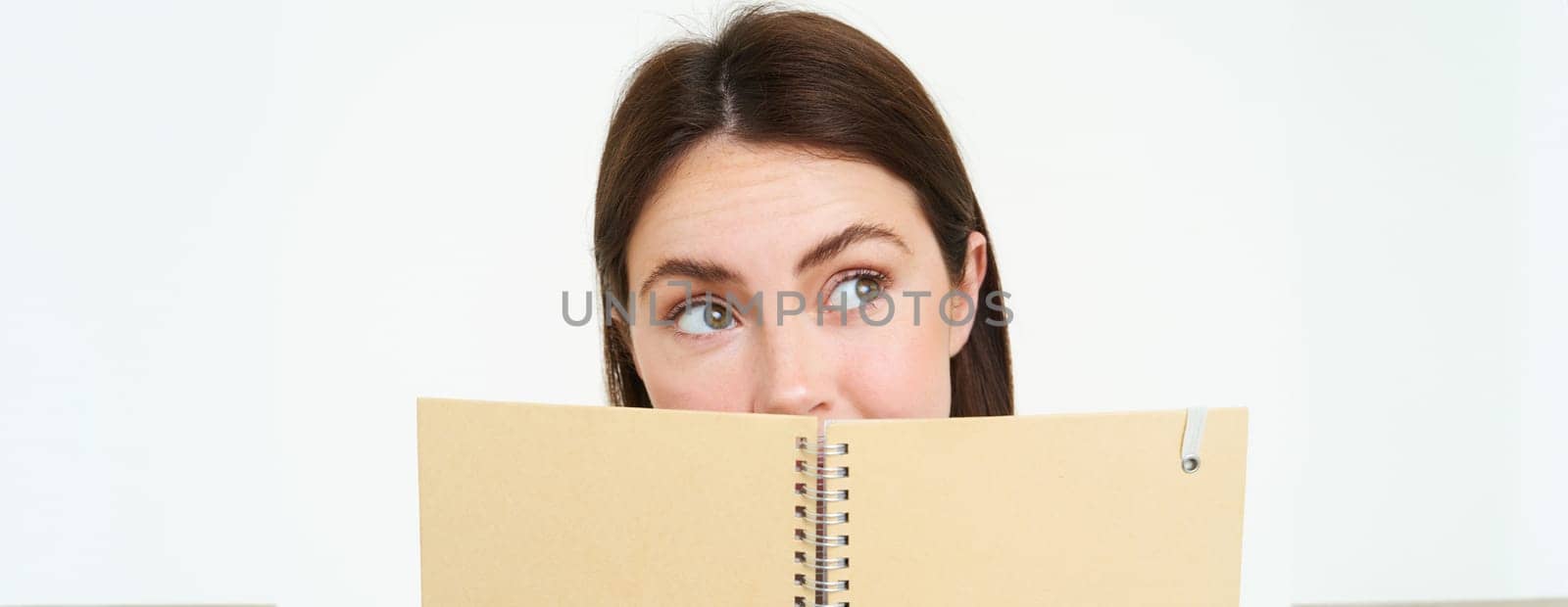 Young woman holds notebook daily planner next to her face, writing down homework, making notes, looking thoughtful, standing over white background by Benzoix