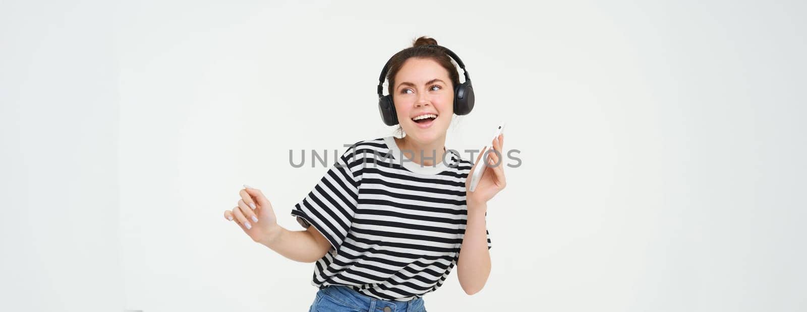 Portrait of stylish young woman dancing in wireless earphones, using headphones and mobile phone music app to listen to favourite songs, posing over white background.