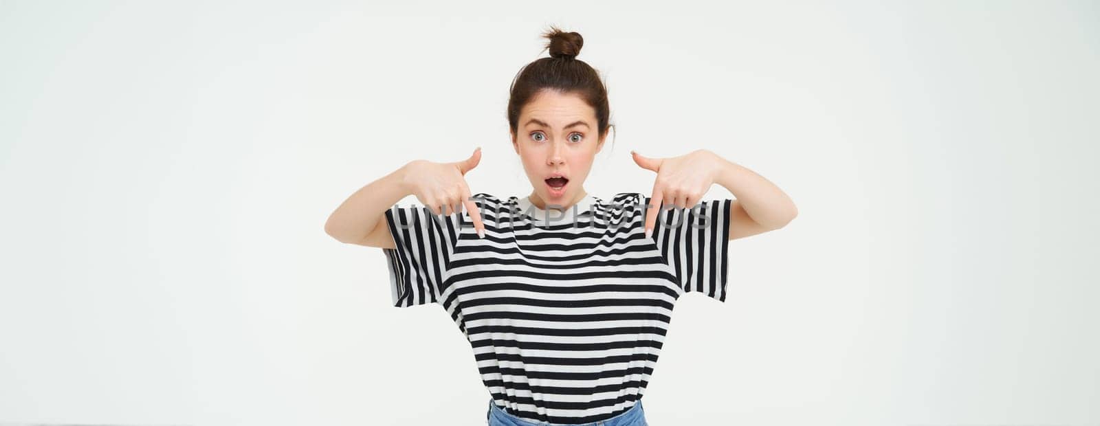 Image of impressed girl, drops jaw, says wow, points fingers down, shows advertisement, being amazed by awesome promo offer, white background.