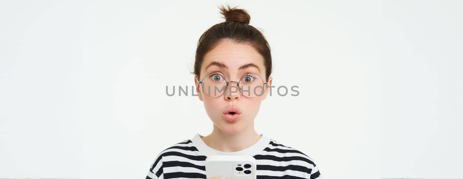 Image of girl in glasses, holding smartphone and looking surprised, amazed by smth on mobile phone, stands over white background.