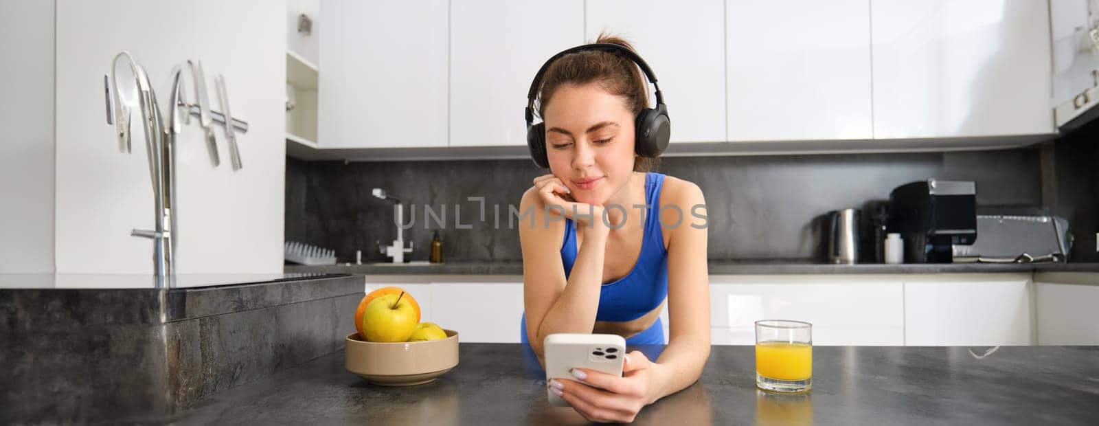 Portrait of young fitness woman with headphones, drinking orange juice in kitchen and using smartphone, listening music, getting ready for workout gym.