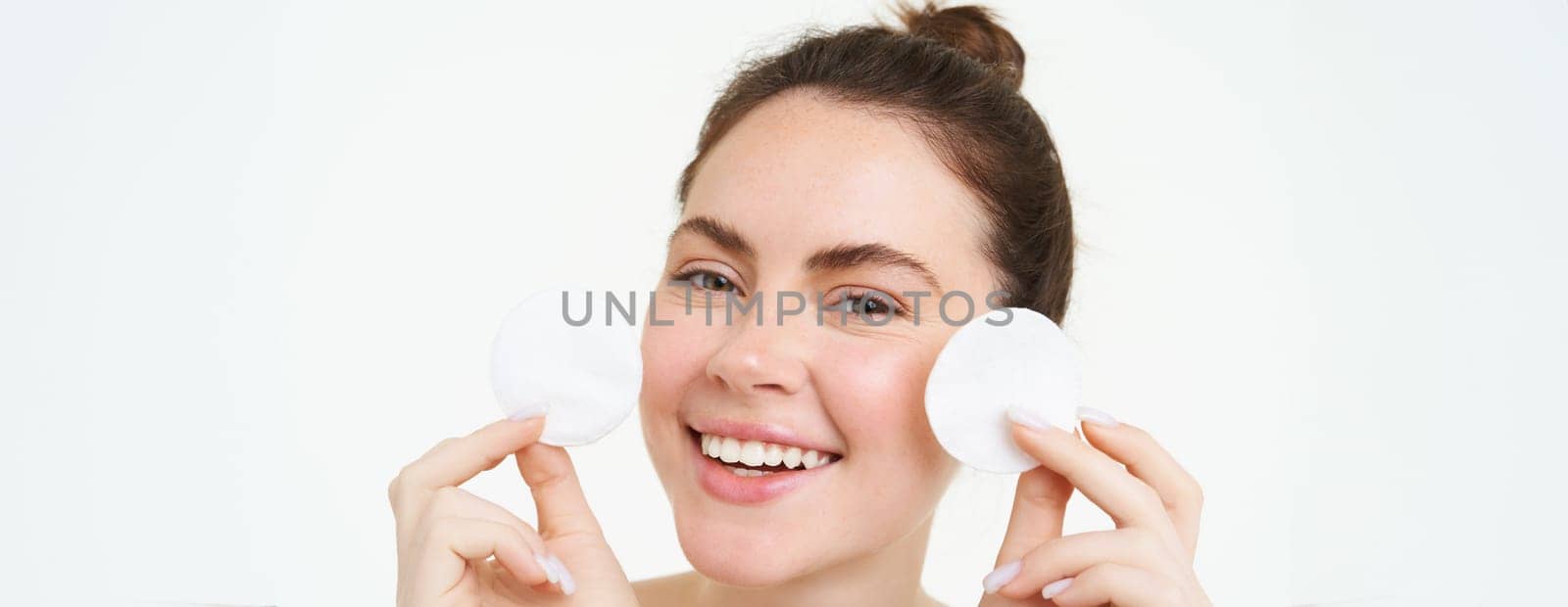 Beauty woman with clear glowing face, showing cotton cosmetic pads for makeup removal, cleansing her facial skin, standing over white background by Benzoix