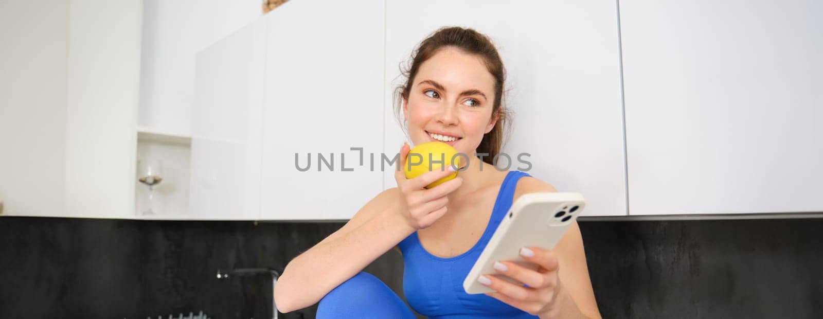 Portrait of sportswoman, girl eating and apple and looking at her social media, smartphone screen, having a snack in kitchen, wearing fitness activewear.