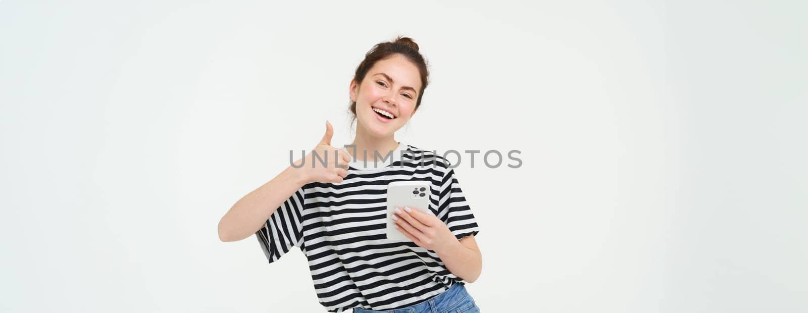 Enthusiastic girl with smartphone shows thumbs up, isolated over white background.