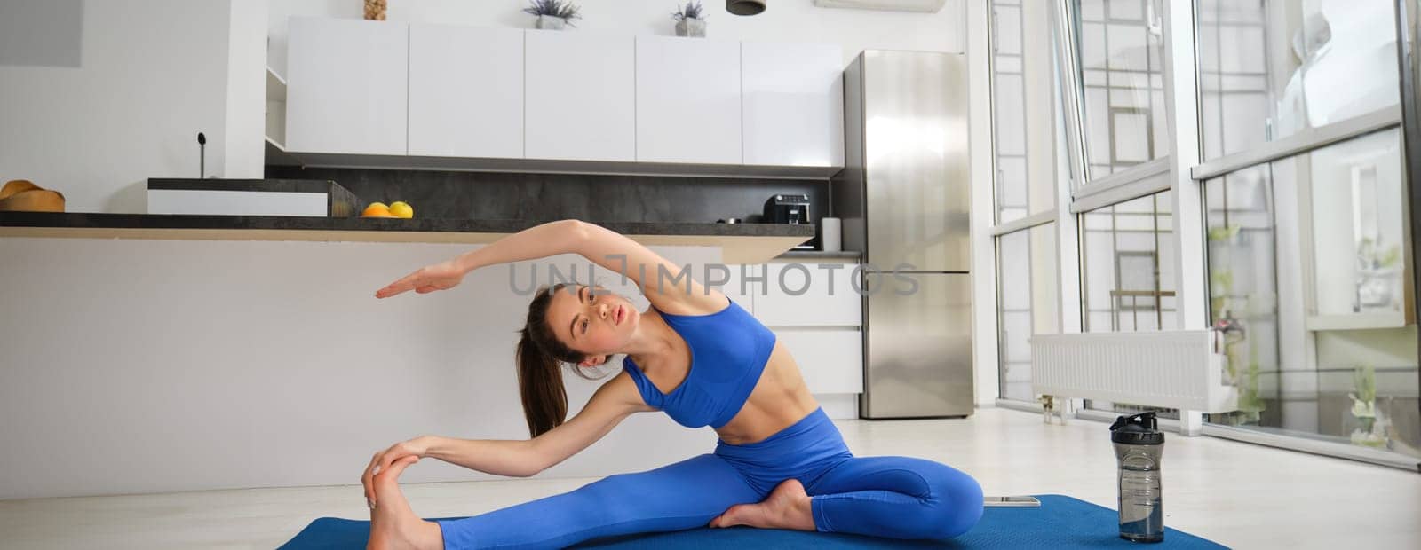 Image of young woman sitting near kitchen on fitness mat, doing yoga exercises, stretching her body, workout from home by Benzoix