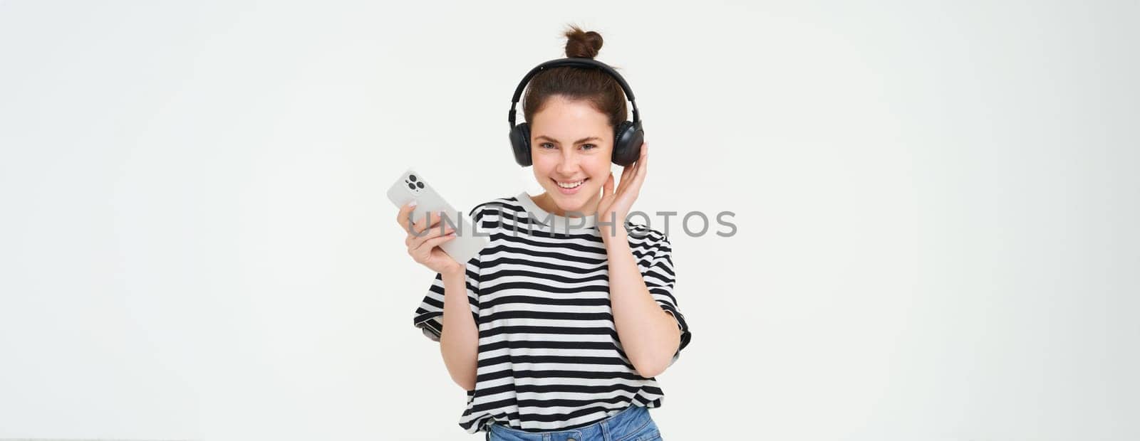 Young woman with smartphone listening to music, dancing to her favourite song in headphones, posing against white background by Benzoix