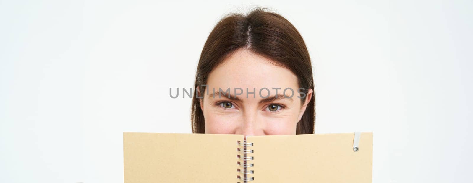 Portrait of cute young woman hides her face behind planner, holds notebook against her face and smiles, isolated over white background.