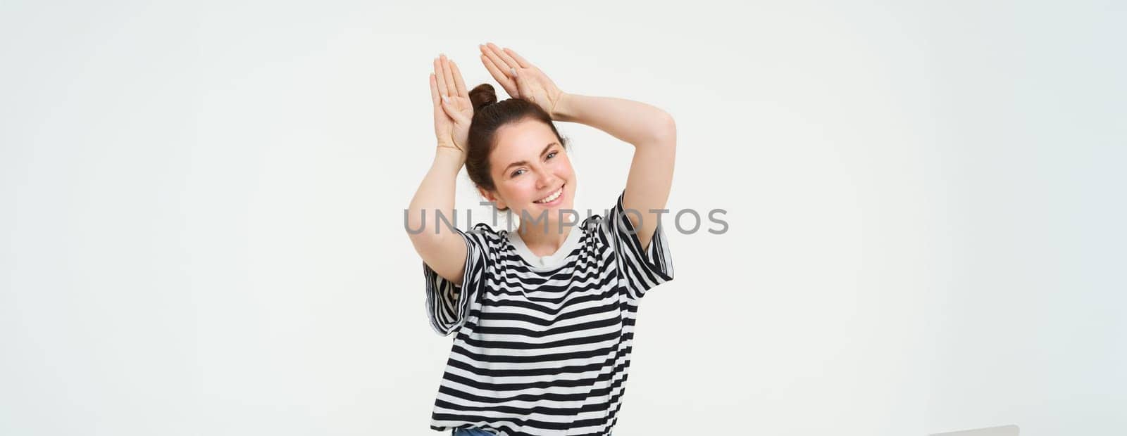 Cute young woman shows animal ears gesture above head, smiling and looking happy, posing in casual clothes over white background by Benzoix