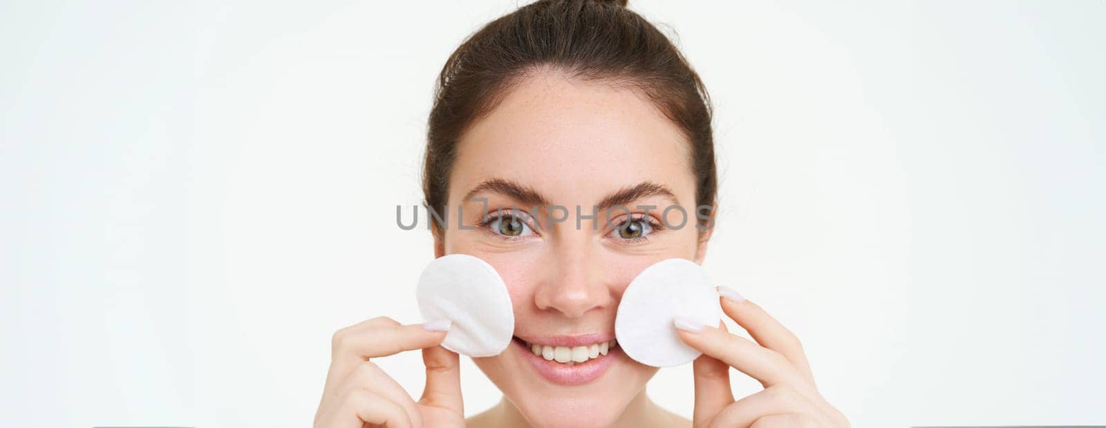 Close-up portrait of young woman washing her face, takes off her makeup with cotton pads, using facial toner, skincare cleanser, standing over white background.