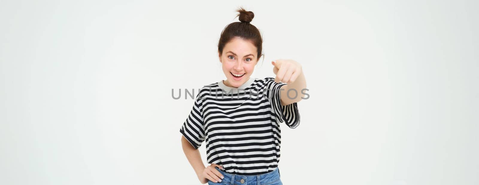 Image of confident, happy young woman in casual clothes, pointing finger at camera, laughing and smiling, standing over white background.