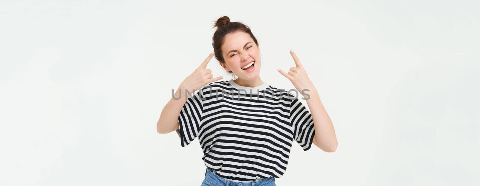 Portrait of excited young woman having fun, rock n roll gesture, enjoys concert, posing upbeat, standing over white background.