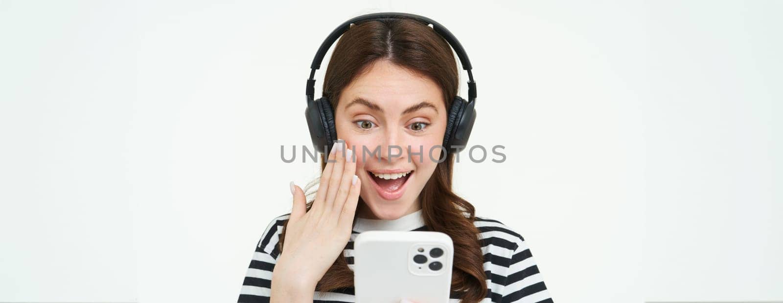 Portrait of beautiful young woman listens to music in wireless headphones, uses her mobile phone, smiles and looks happy, isolated on white background.