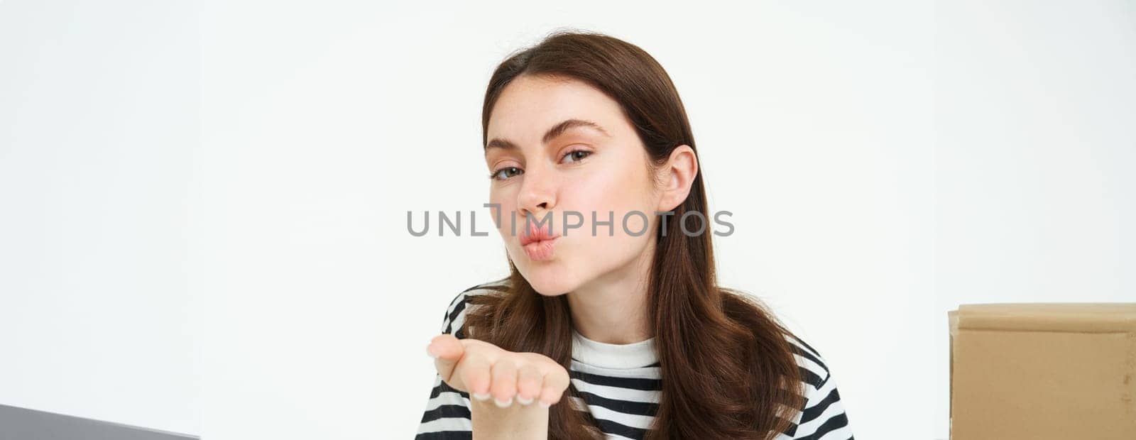 Portrait of cute girl sending you, blowing air kiss, holding palm near puckered lips, smiling, standing over white background.
