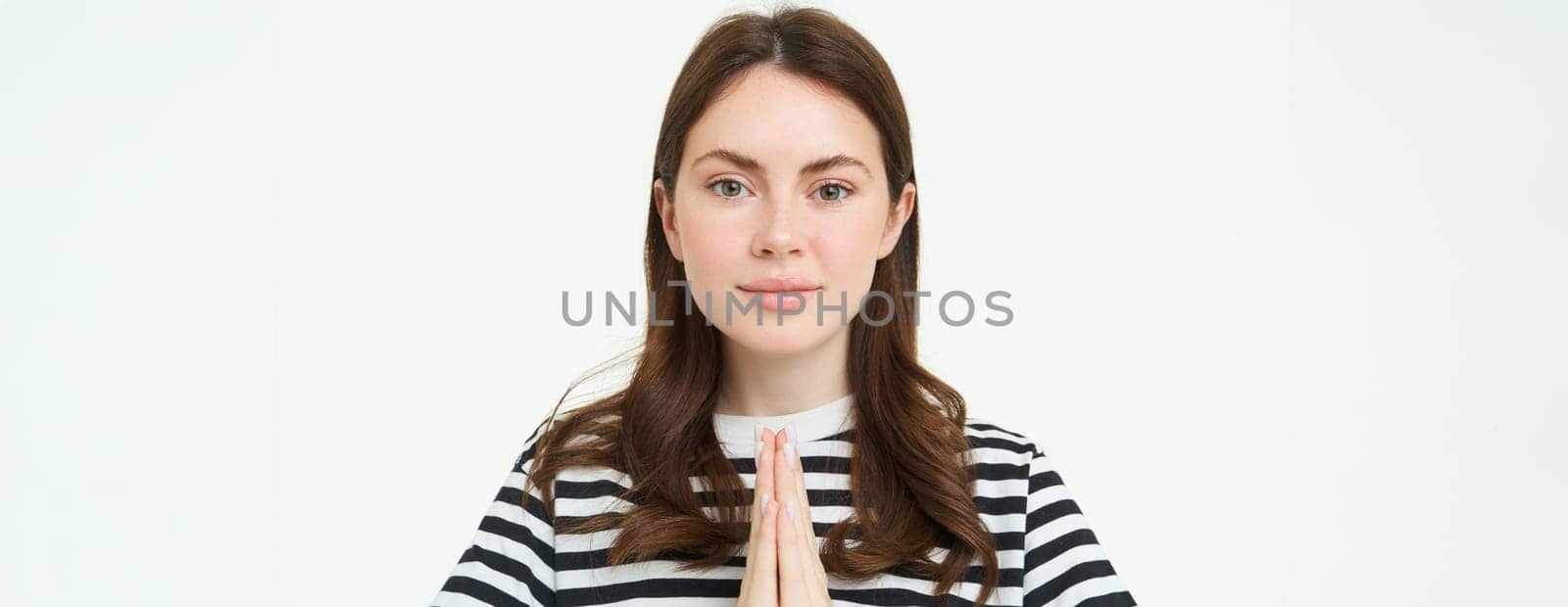 Portrait of young woman expresses her gratitude, shows thank you, namaste gesture, holding hands clasped together near chest and smiling, standing over white background.