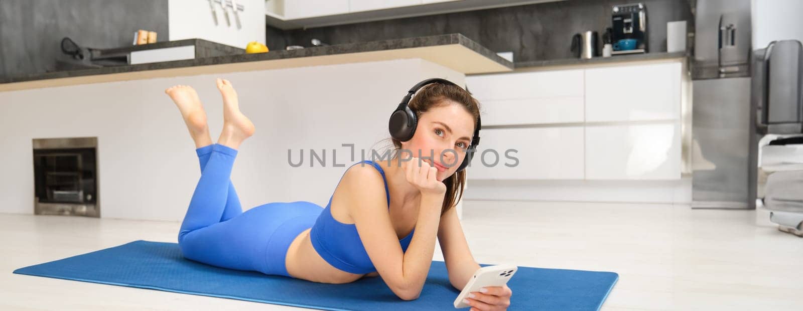 Portrait of beautiful fitness girl, laying on yoga mat, using mobile phone and wireless headphones, looking thoughtful and smiling.